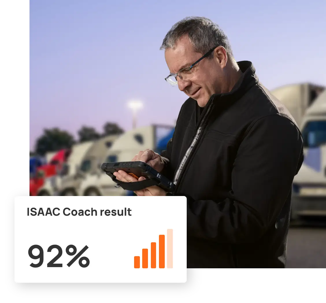 Man smiling while looking at a handheld device displaying 'ISAAC Coach result 92%' with a bar graph, standing in a parking lot with trucks in the background.