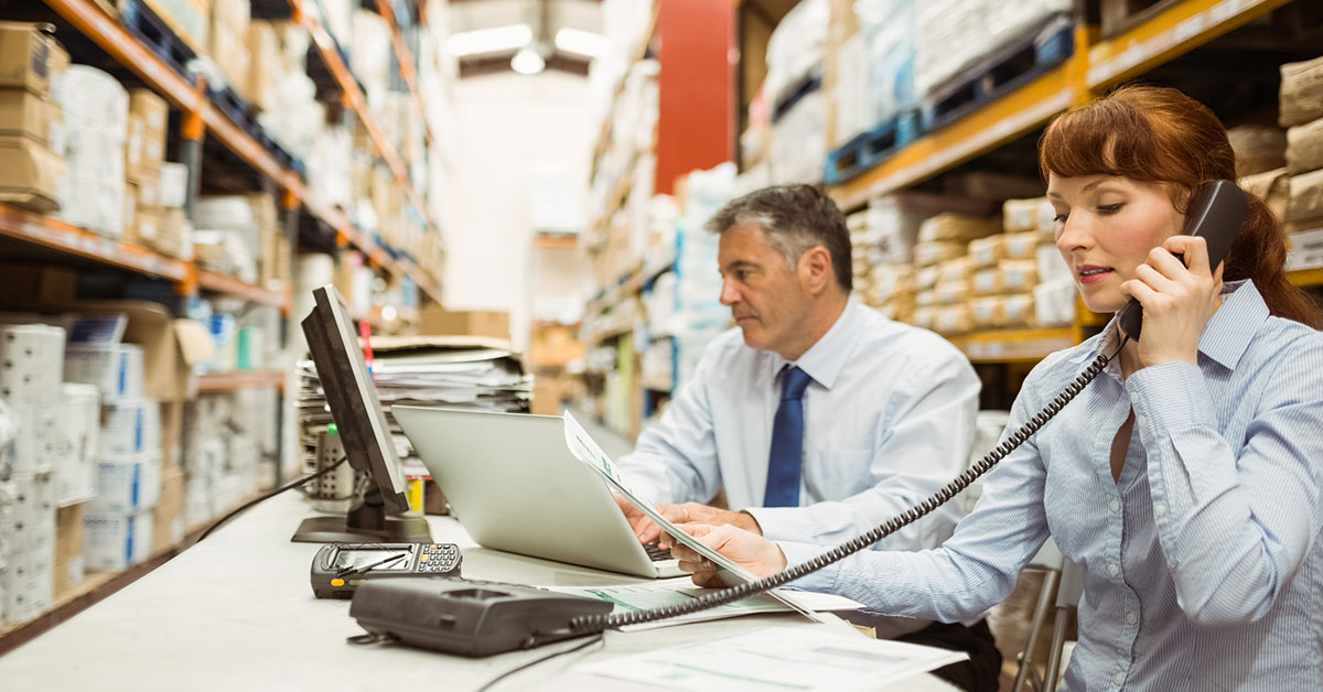 Two employees using phones and computers within a warehouse, showing internal company communication supporting customer service goals.