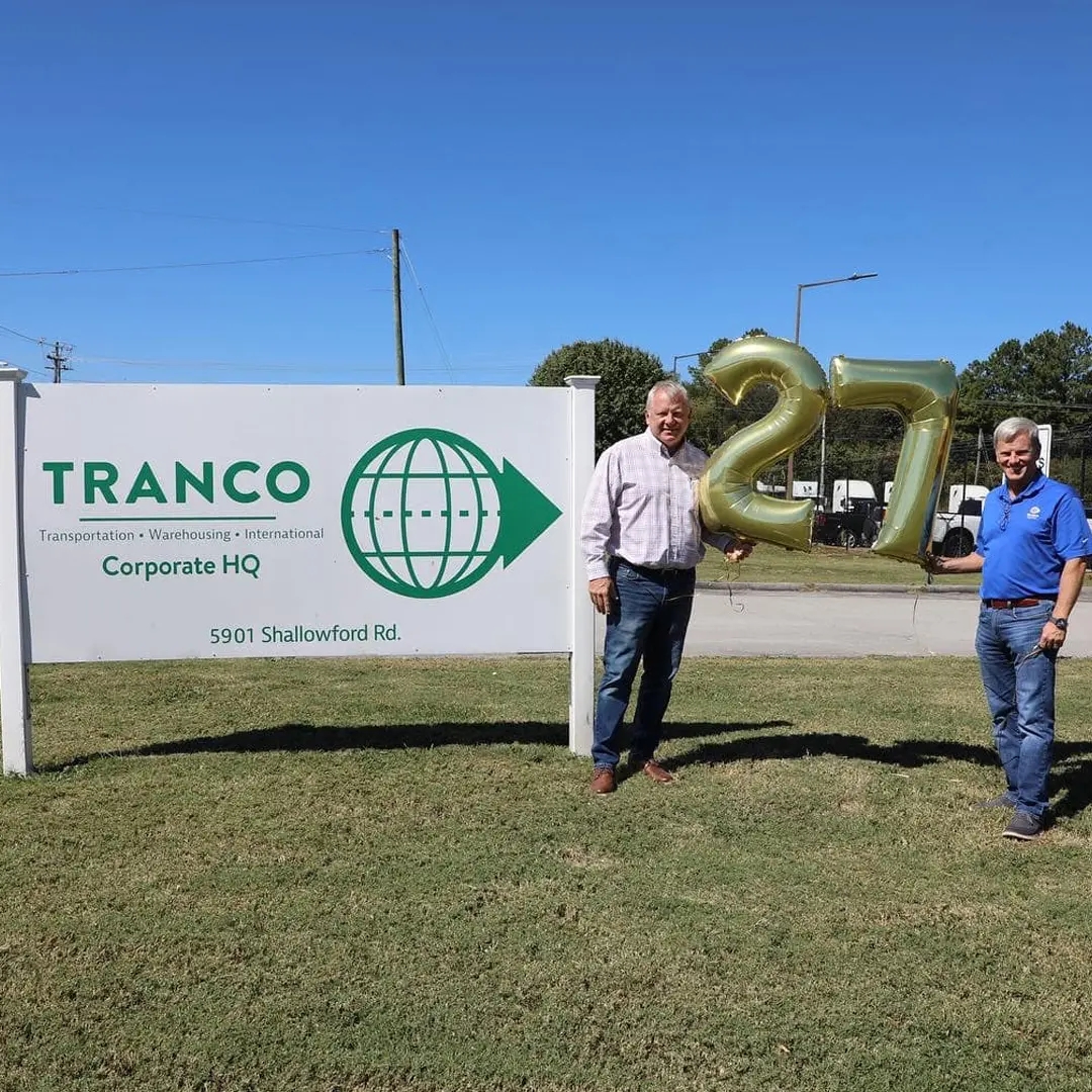 Two men holding gold “27” balloons beside the Tranco Logistics Corporate HQ sign.