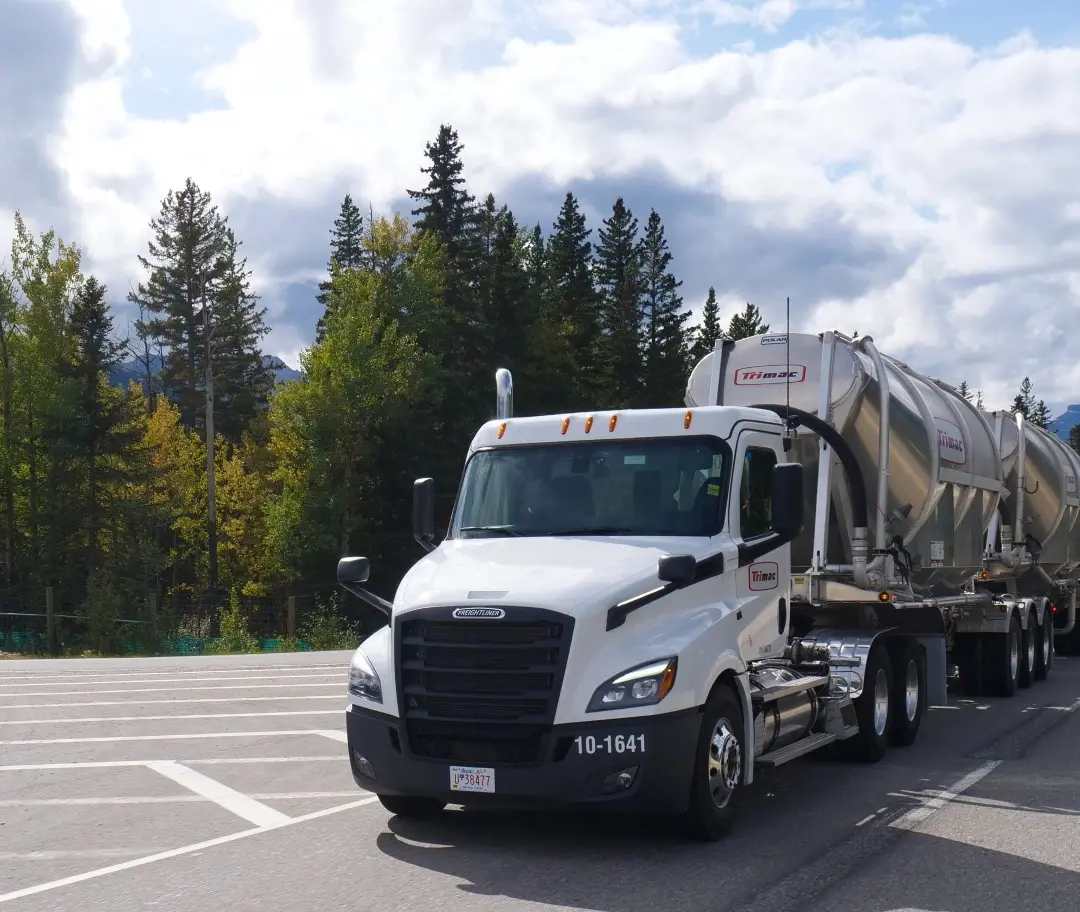 A Trimac bulk tanker truck parked roadside with lush green trees in the background under a cloudy sky.