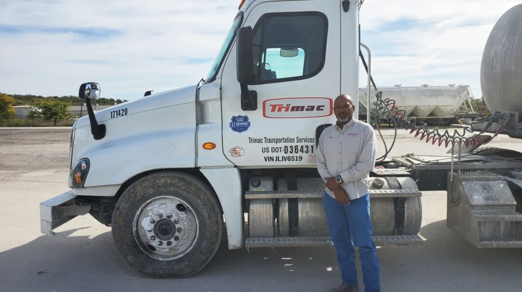 A Trimac truck driver standing proudly beside a company truck, displaying the Trimac and ISAAC logos.