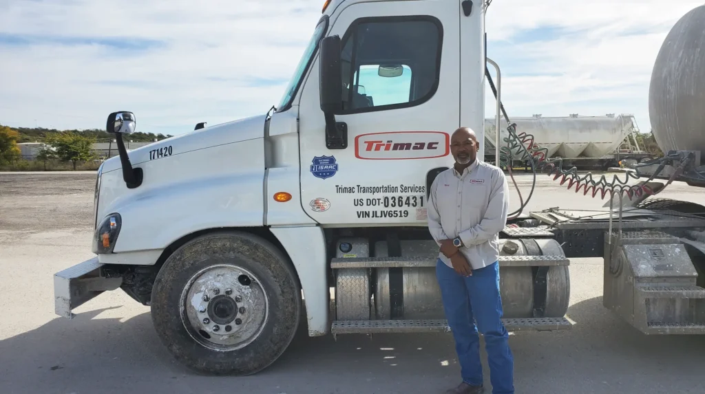 Trimac Professional Driver and Truck A Trimac truck driver standing proudly beside a company truck, displaying the Trimac and ISAAC logos.