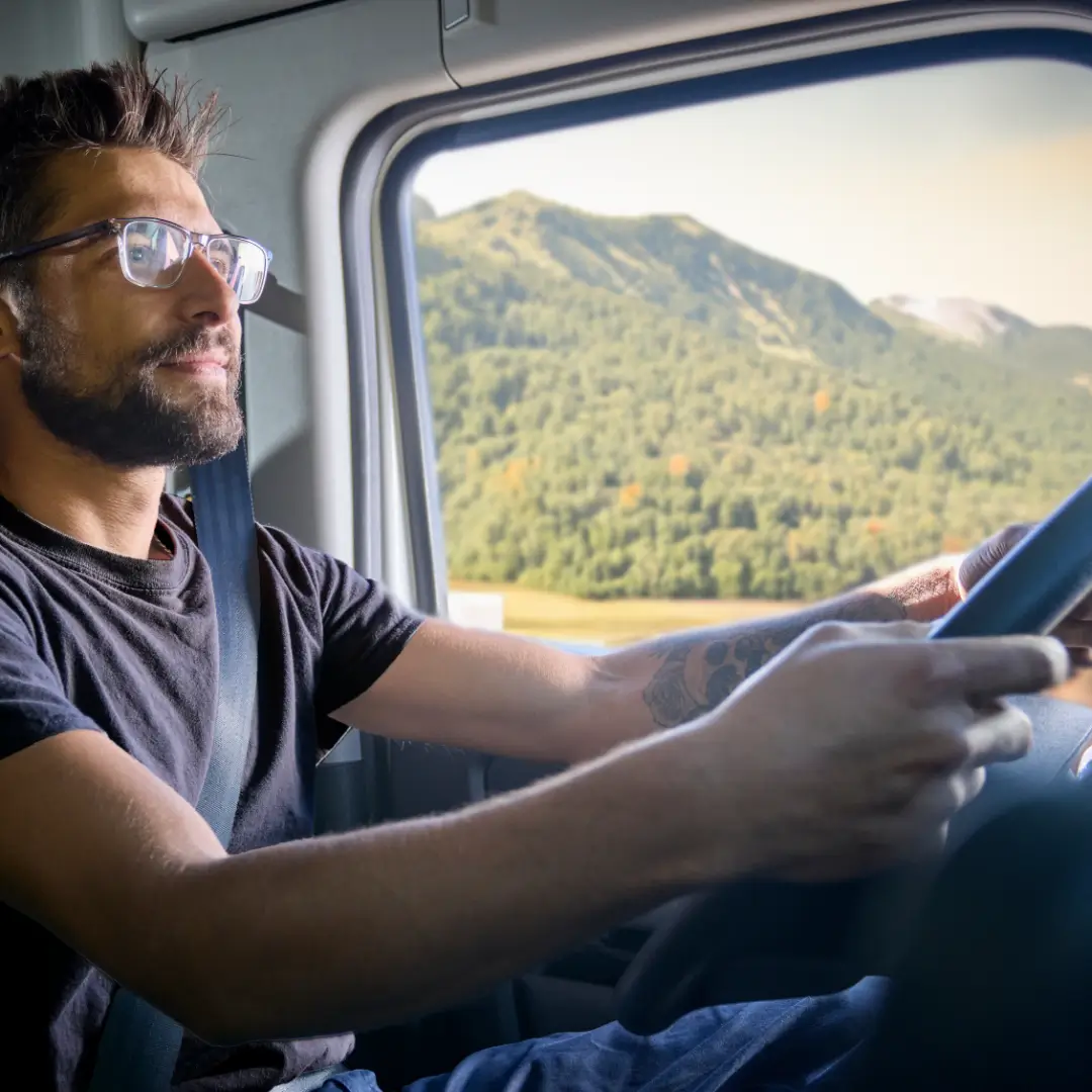 Truck Driver Navigating Mountain Roads Male truck driver with glasses driving a truck with a scenic mountain view in the background.