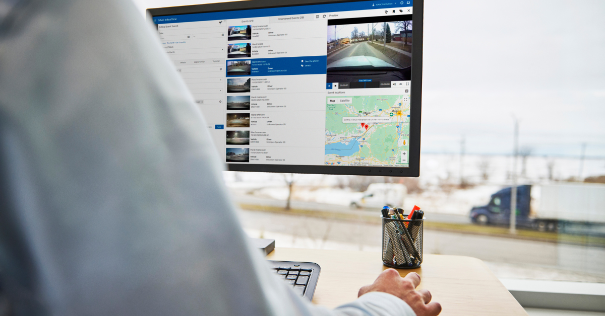 Fleet management software dashboard showing real-time vehicle tracking data with location map and video feed, displayed on a monitor in an office with a blurred highway view in the background.