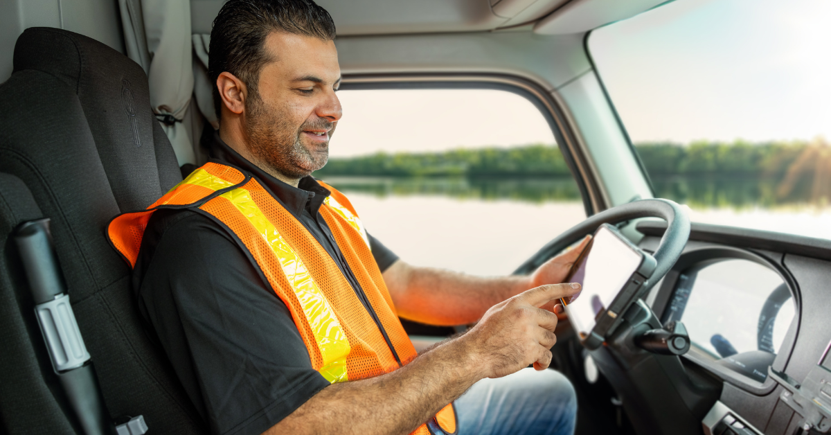 A truck driver wearing an orange safety vest using an ISAAC ELD device in a truck near a scenic river.