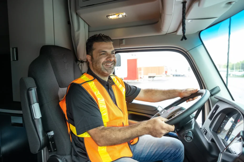 A truck driver in an orange safety vest sitting in the driver’s seat of a truck, smiling.