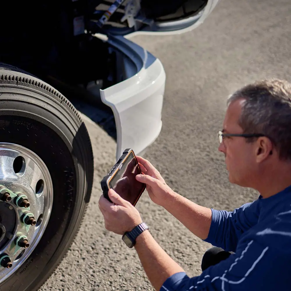 Driver using ISAAC device to record pre-trip inspection by truck wheel