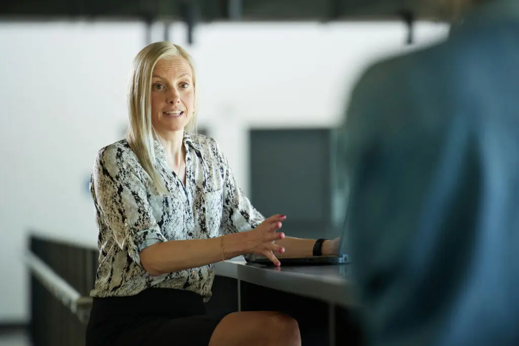 Businesswoman in conversation during a professional check-in meeting, sitting at a table with a laptop